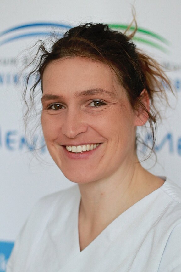 Nahaufnahme einer lächelnden Person in weißer medizinischer Kleidung, Blick in die Kamera. Haare locker zurückgebunden. Heller, unscharfer Hintergrund.