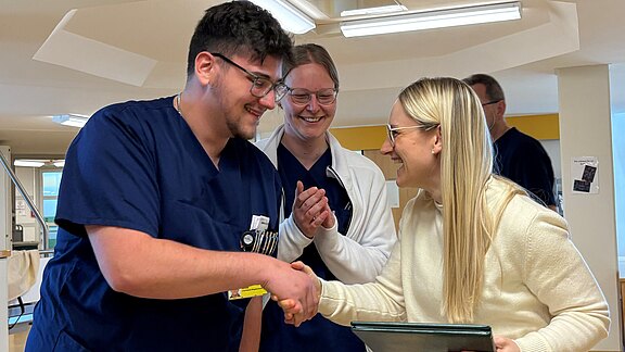 In einem Krankenhaus schüttelt eine Frau mit Brille und Tablet einem Pfleger in blauer Dienstkleidung die Hand; zwei Kollegen im Hintergrund lächeln und klatschen. Szene wirkt wie Gratulation oder Anerkennung.