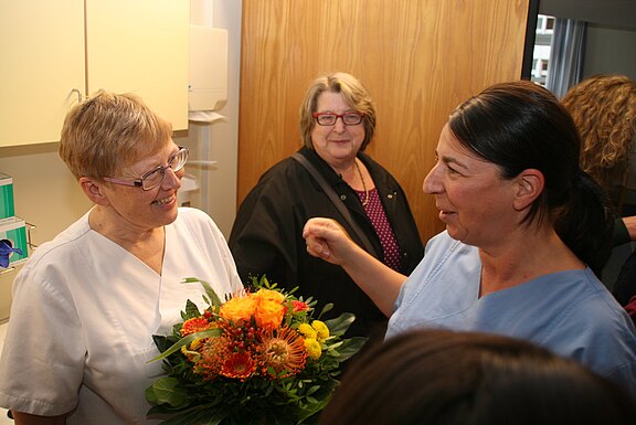 Zwei Pflegekräfte in einem Behandlungsraum unterhalten sich lächelnd. Eine hält einen Strauß orange-gelber Blumen, offenbar als Gratulation oder Abschied. Weitere Personen stehen daneben und beobachten die Szene.