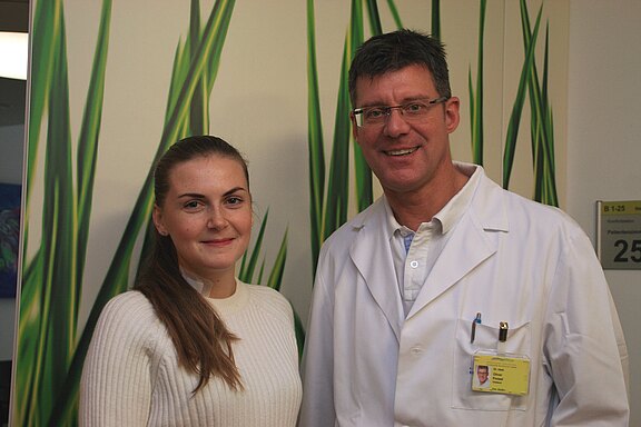 Zwei Personen stehen nebeneinander und lächeln in die Kamera: links eine Frau im hellen Pullover, rechts ein Mann im weißen Kittel mit Namensschild. Der Hintergrund zeigt eine Wand mit grünen Pflanzenmotiven, wirkt wie ein Klinikflur.