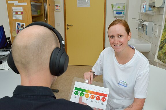 Eine Frau lächelt und zeigt einer Person ein Bewertungsblatt mit Smiley-Gesichtern, das zur Einschätzung des Hörvermögens dient. Die Person trägt Kopfhörer und sitzt an einem Tisch in einer Praxis. Im Hintergrund sind medizinische Geräte erkennbar.