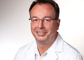 Porträt eines Mannes mittleren Alters mit Brille im weißen Kittel. Er lächelt freundlich und blickt in die Kamera vor neutral hellem Hintergrund.