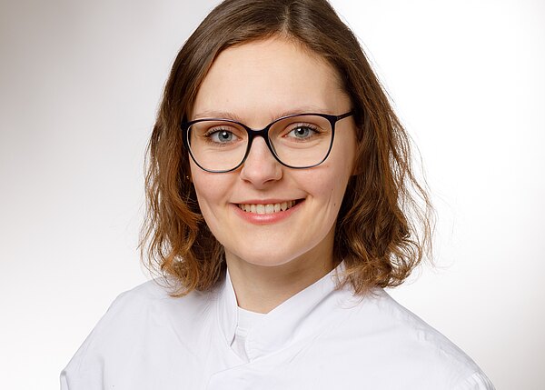 Porträt einer jungen Frau mit schulterlangen braunen Haaren und schwarzer Brille, die lächelt. Sie trägt einen weißen Kittel vor neutralem Hintergrund.