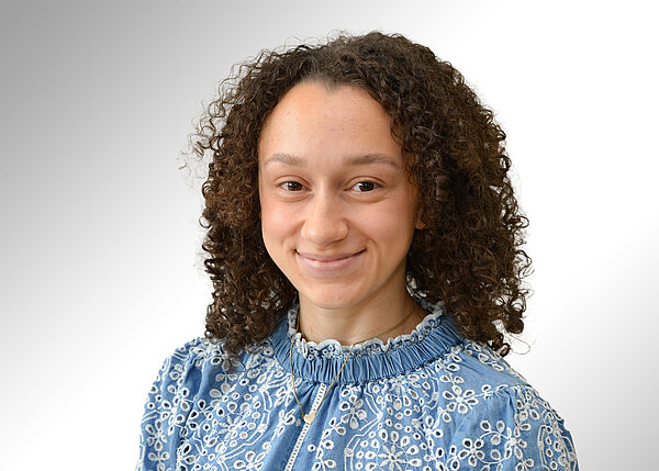 Porträt einer lächelnden Person mit dunkelbraunen Locken, leicht nach rechts gewandt. Sie trägt eine hellblaue, reich bestickte Bluse mit hohem Kragen. Neutraler, heller Hintergrund.