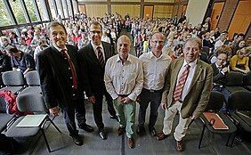Eine Gruppe von fünf Männern steht im Vordergrund, während im Hintergrund eine große Menschenmenge in einem Raum sitzt. Die Atmosphäre wirkt informell und lebhaft, was auf eine Veranstaltung oder eine Ansprache hindeutet.