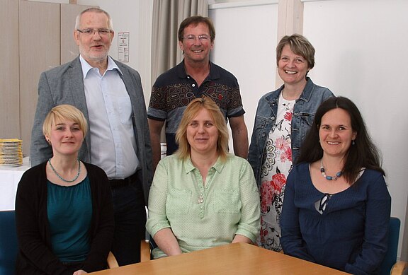 Gruppenfoto: sechs Erwachsene in einem hellen Raum. Drei sitzen an einem Holztisch, drei stehen dahinter. Alle schauen in die Kamera und lächeln. Informelle, berufliche Atmosphäre.
