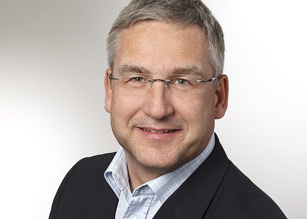 Porträtfoto: Ein grauhaariger Mann mittleren Alters mit randloser Brille lächelt. Er trägt ein dunkles Sakko und ein hellblau kariertes Hemd. Neutraler, heller Hintergrund.