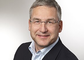 Porträtfoto: Ein grauhaariger Mann mittleren Alters mit randloser Brille lächelt. Er trägt ein dunkles Sakko und ein hellblau kariertes Hemd. Neutraler, heller Hintergrund.