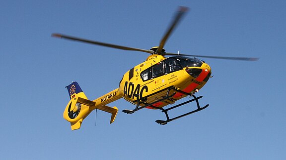 Gelber Rettungshubschrauber mit der Aufschrift „ADAC“ und „Notarzt“ fliegt vor blauem Himmel; Rotoren in Bewegung, Landekufen sichtbar.