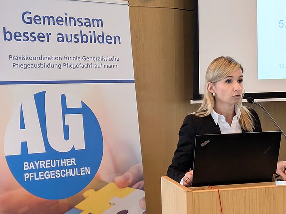 Eine Frau hält einen Vortrag an einem Rednerpult mit Mikrofon und Laptop. Neben ihr steht ein Roll‑Up mit der Aufschrift „Gemeinsam besser ausbilden“ und dem Logo „AG Bayreuther Pflegeschulen“. Thema: Praxiskoordination und generalistische Pflegeausbildung.