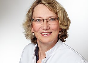 Porträt einer lächelnden Frau mit Kurzhaar und Brille vor neutralem Hintergrund. Sie trägt einen weißen Kittel mit offenem Kragen und Perlenohrringe; freundlicher, direkter Blick in die Kamera.