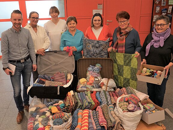 Sieben Personen stehen hinter einem Tisch und zeigen zahlreiche handgestrickte Socken, Mützen und Decken. Die bunten Spenden liegen geordnet in offenen Koffern, Taschen und Kisten.
