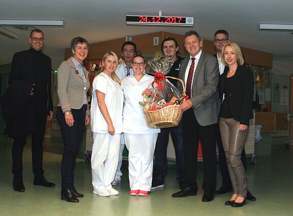Neun Erwachsene posieren in einem Krankenhausflur; zwei Personen in weißer Pflegekleidung stehen in der Mitte. Eine Person überreicht einen großen, weihnachtlich dekorierten Geschenkkorb. Die Gruppe lächelt in die Kamera; es wirkt wie ein Dankeschön an das Pflegepersonal.