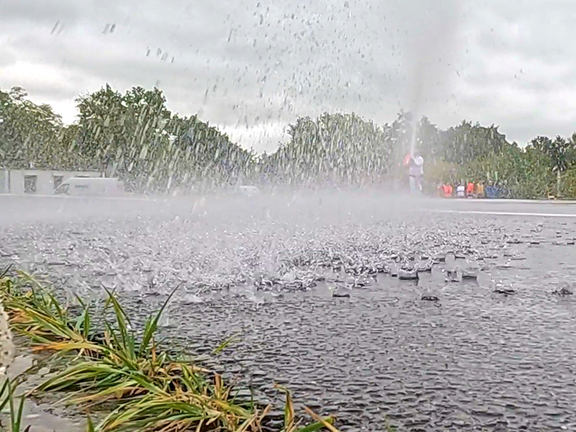 Ein Wasserstrahl spritzt aus dem Boden, während sich der Regen auf der asphaltierenden Fläche verteilt. Im Hintergrund sind grüne Bäume und verschwommene Figuren zu erkennen, die sich in einer leicht nebligen Umgebung bewegen. Der Himmel ist bewölkt.