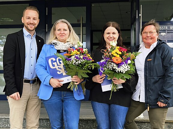 Vier Personen stehen auf einer Treppe vor einem Gebäude. Zwei Frauen halten bunte Blumensträuße, während ein Mann und eine Frau lächeln. Alle wirken erfreut und stehen in einer geselligen Atmosphäre.