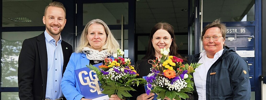 Vier Personen stehen auf einer Treppe vor einem Gebäude. Zwei Frauen halten bunte Blumensträuße, während ein Mann und eine Frau lächeln. Alle wirken erfreut und stehen in einer geselligen Atmosphäre.