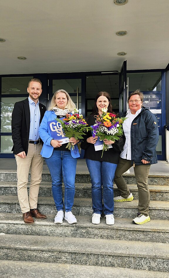 Vier Personen stehen auf einer Treppe vor einem Gebäude. Zwei Frauen halten bunte Blumensträuße, während ein Mann und eine Frau lächeln. Alle wirken erfreut und stehen in einer geselligen Atmosphäre.