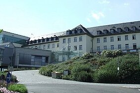 Ein mehrstöckiges Gebäude mit moderner Glasarchitektur ist im Vordergrund zu sehen. Im Hintergrund ist ein traditionellerer Gebäudeteil mit einem Satteldach. Um das Gebäude herum gibt es eine grüne Landschaft mit Blumen und Büschen.