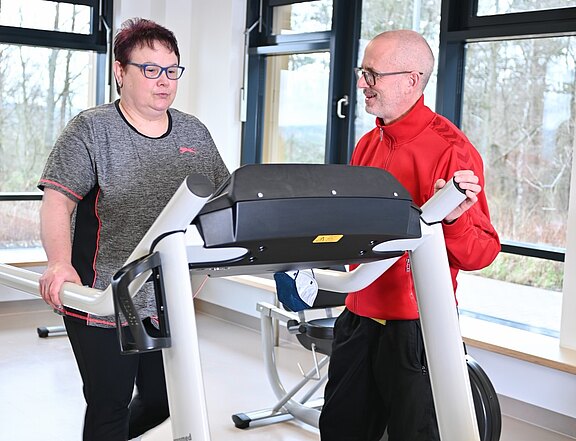 In einem hellen Fitnessraum trainiert eine Frau auf einem Laufband. Ein Trainer steht neben ihr, spricht mit ihr und kontrolliert die Einstellungen. Die Szene zeigt betreutes Ausdauertraining.