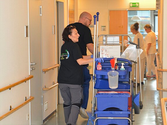 Zwei Reinigungskräfte schieben einen Reinigungswagen durch einen Krankenhausflur. Auf dem Wagen stehen Eimer, Tücher und Putzmittel. Im Hintergrund gehen Pflegekräfte den Gang entlang. Die Szene zeigt Gebäudereinigung im Klinikalltag.