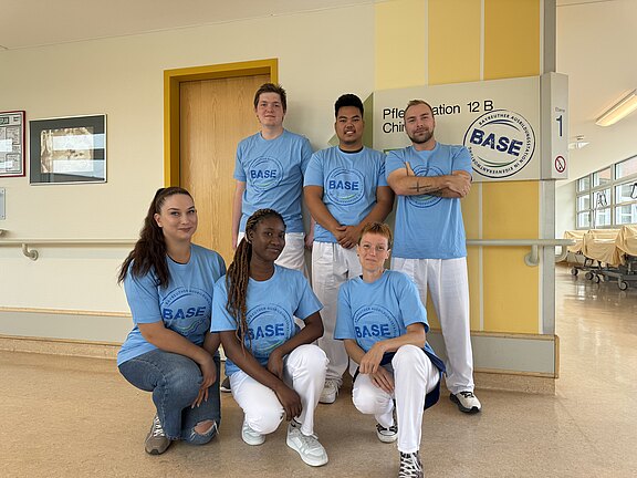 Sechs junge Erwachsene in hellblauen BASE-T-Shirts, teils in weißer Arbeitskleidung, posieren im Flur einer Krankenhausstation. Drei stehen, drei hocken davor. Im Hintergrund ein Stationsschild. Gruppenfoto eines Pflege- oder Ausbildungsteams auf einer Station.