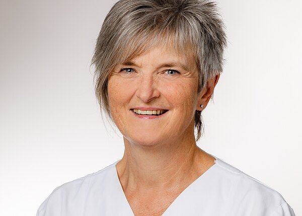 Eine lächelnde Frau mit kurzen, grauen Haaren trägt eine weiße Berufsbekleidung. Sie hat einen freundlichen Gesichtsausdruck und schaut direkt in die Kamera. Der Hintergrund ist einfarbig und erzeugt einen professionellen Eindruck.