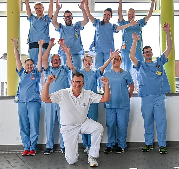 Eine Gruppe von zehn medizinischen Mitarbeitenden in blauen Kitteln, mit einer Person in Weiß vorn kniend, jubelt und hebt die Arme; alle lächeln in einem hellen Flur.