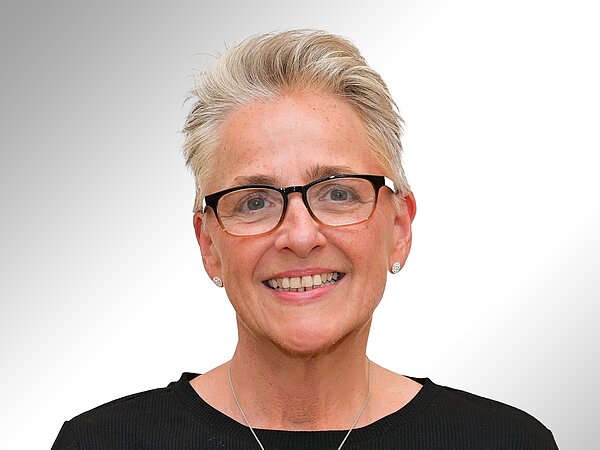 Porträt einer lächelnden Frau mit kurzen, grauen Haaren und Brille. Sie trägt ein schwarzes Oberteil und Ohrstecker. Der Hintergrund ist hell und weich verschwommen.