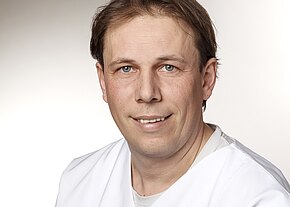 Porträt eines Mannes in weißer Arbeitskleidung vor neutralem Hintergrund, der direkt in die Kamera blickt und leicht lächelt.