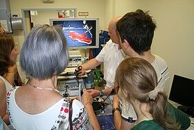 Eine Gruppe von Menschen steht um einen Arzt, der ihnen ein medizinisches Bild auf einem Monitor zeigt. Der Arzt erklärt etwas, während die anderen interessiert zuhören und einige Haltegeräte in den Händen halten.
