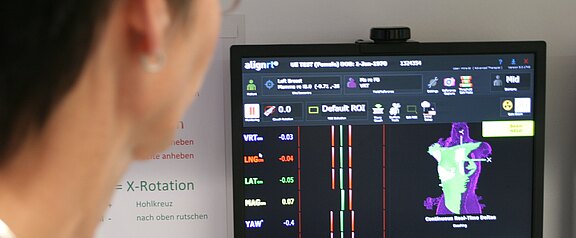 Eine Person betrachtet einen Monitor, der eine Software zur Haltungsanalyse zeigt: farbige Balken mit Messwerten und eine 3D-Silhouette eines Oberkörpers. Die Darstellung verdeutlicht Abweichungen und Ausrichtung von Wirbelsäule und Körperachsen.