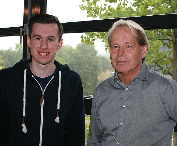 Zwei Männer stehen nebeneinander in einem Innenraum vor Fenstern. Links ein junger Mann im dunklen Hoodie, lächelnd. Rechts ein älterer Mann im grauen Hemd, ernster Blick; beide schauen in die Kamera.