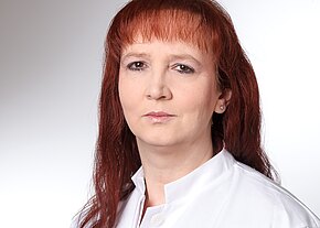 Nahaufnahme einer erwachsenen Person mit rötlichen, schulterlangen Haaren und neutralem Blick. Sie trägt einen weißen Kittel vor hellem, neutralem Hintergrund.