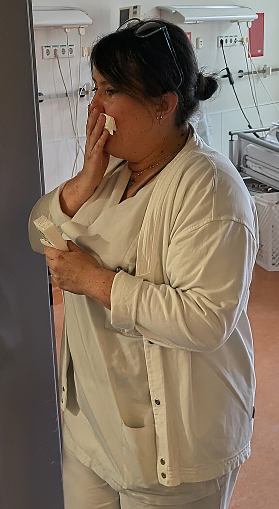 Eine Pflegekraft in weißer Arbeitskleidung steht in einem Krankenhausraum, hält ein Taschentuch an Mund und Nase und ein weiteres in der Hand. Sie wirkt nachdenklich und erschöpft.