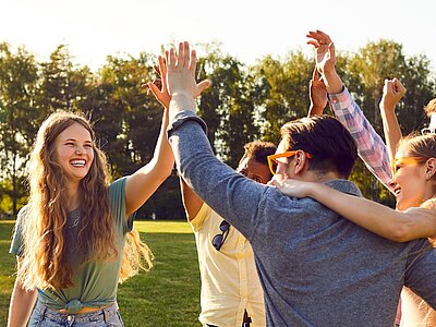 Gruppe junger Menschen in einem Park feiert lachend; mehrere heben die Arme und geben High-Fives. Sonnenlicht, sommerliche Stimmung, gemeinschaftliche Freude auf einer Wiese.