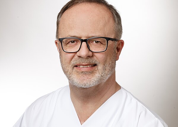 Porträt eines Mannes mittleren Alters mit Brille und grauem Bart, in weißer medizinischer Kleidung, vor neutralem Hintergrund, mit freundlichem Ausdruck.