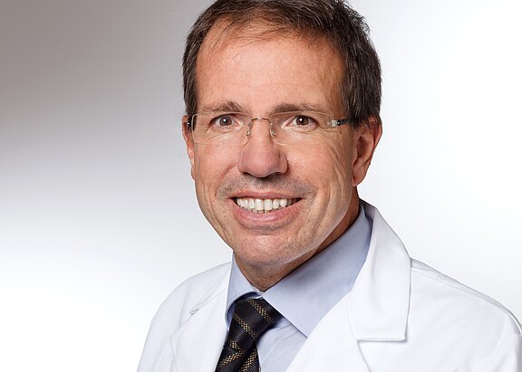 Porträt eines lächelnden Mannes mittleren Alters mit Brille, im weißen Arztkittel, blauem Hemd und dunkler Krawatte vor neutralem Hintergrund.