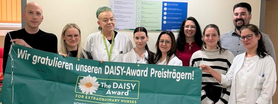 Gruppe von Pflegekräften und Kolleginnen/Kollegen im Krankenhausflur hält ein großes grünes Banner mit der Aufschrift: „Wir gratulieren unseren DAISY-Award Preisträgern! The DAISY Award for Extraordinary Nurses.“ Feierliche Anerkennung für besondere Pflegeleistungen.