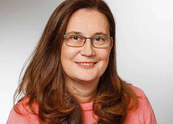 Porträt einer erwachsenen Frau mit langen braunen Haaren und Brille. Sie lächelt freundlich in die Kamera und trägt einen rosa Pullover vor neutral hellem Hintergrund.