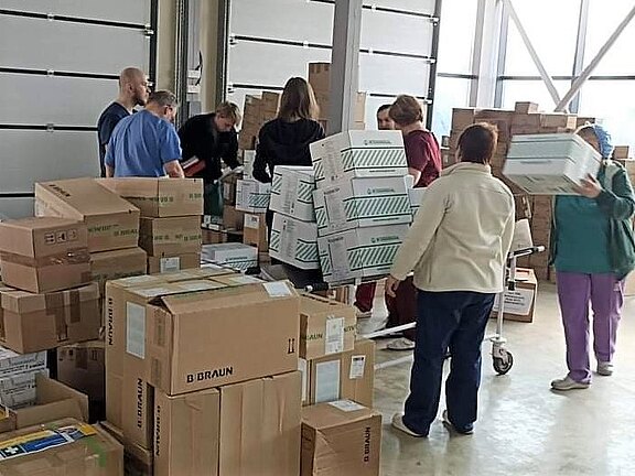 Mehrere Personen arbeiten in einer Halle zwischen vielen Kartons. Sie stapeln und sortieren Kisten und laden sie auf Rollwagen.