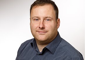 Halbnahes Porträt eines leicht lächelnden Mannes. Kurze braune Haare, dezenter Bart. Er trägt ein dunkelgraues Hemd mit offenem Kragen. Heller, neutraler Hintergrund.