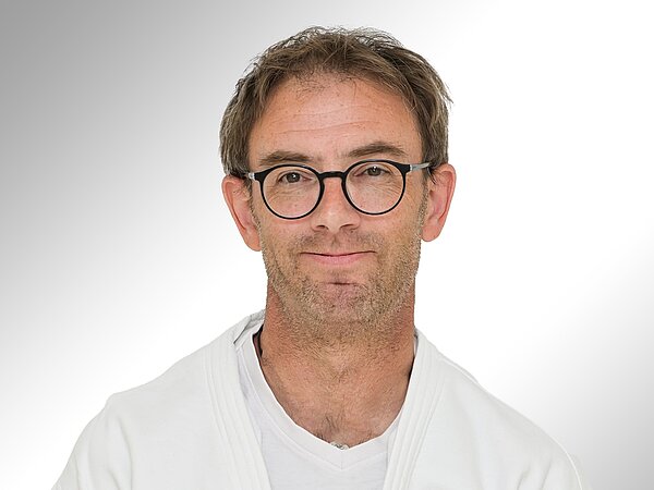 Ein Mann mit Brille und kurzem, leicht zerzaustem Haar trägt einen weißen Judo-Anzug. Er lächelt freundlich in die Kamera. Der Hintergrund ist hell und neutral gehalten, was den Fokus auf sein Gesicht lenkt.