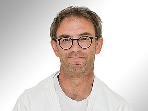 Ein Mann mit Brille und kurzem, leicht zerzaustem Haar trägt einen weißen Judo-Anzug. Er lächelt freundlich in die Kamera. Der Hintergrund ist hell und neutral gehalten, was den Fokus auf sein Gesicht lenkt.