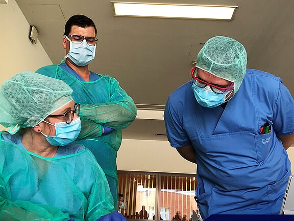 Im Bild sind drei medizinische Fachkräfte in Schutzausrüstung zu sehen. Zwei Personen stehen und schauen auf eine dritte Person, die sitzt und etwas untersucht. Alle tragen Masken und Schutzbrillen. Die Umgebung wirkt klinisch und professionell.