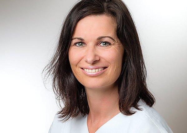 Porträtfoto: Eine erwachsene Frau mit schulterlangem dunklem Haar lächelt in die Kamera. Sie trägt helle Kleidung, neutraler Hintergrund, freundlicher Ausdruck.