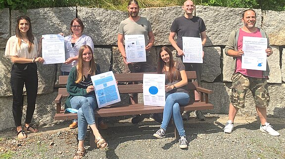 Sieben Personen posieren im Freien vor einer Steinmauer, einige sitzen auf einer Bank. Alle halten große Infoplakate zu medizinischen Themen wie Sepsis, Impfungen und Kalium. Das Bild vermittelt eine Projekt- oder Präsentationssituation.