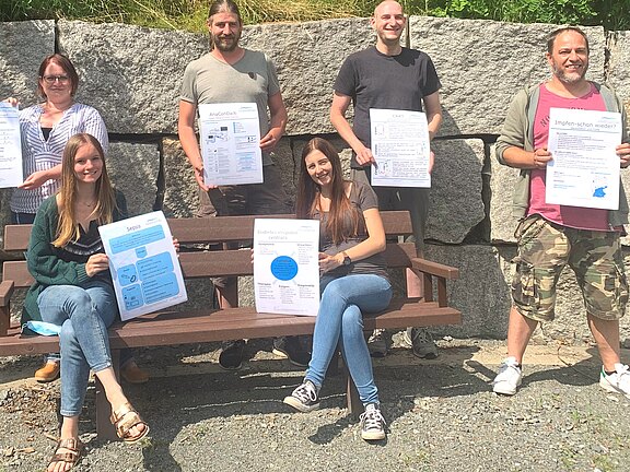 Sieben Personen posieren im Freien vor einer Steinmauer, einige sitzen auf einer Bank. Alle halten große Infoplakate zu medizinischen Themen wie Sepsis, Impfungen und Kalium. Das Bild vermittelt eine Projekt- oder Präsentationssituation.