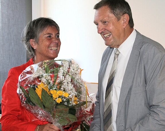 Zwei Erwachsene in formeller Kleidung lächeln. Die Frau in roter Jacke überreicht dem Mann im Anzug einen großen Blumenstrauß mit gelben und weißen Blüten. Die Szene wirkt wie eine Gratulation oder ein Dankesmoment in einem Innenraum.