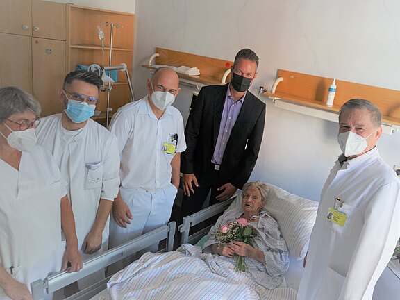Ein älterer Mensch liegt in einem Krankenhausbett und hält einen Blumenstrauß. Um sie herum stehen fünf Personen, darunter medizinisches Personal und ein Mann in Anzug. Alle tragen Masken. Die Atmosphäre ist freundlich und zeigt eine Unterstützungserfahrung im Krankenhaus.