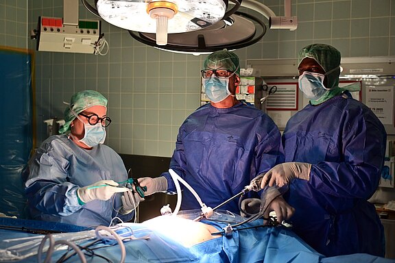 Drei Chirurginnen und Chirurgen in OP-Kleidung führen unter hellem OP-Licht eine minimalinvasive, laparoskopische Operation durch. Sie tragen Masken, Hauben und sterile Handschuhe und bedienen Kameras und Instrumente über kleine Zugänge; der Patient ist abgedeckt, nur das Operationsfeld beleuchtet.